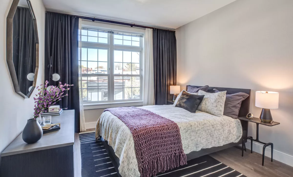 Apartment bedroom with large window, curtains, bed, and furniture at The Bradford