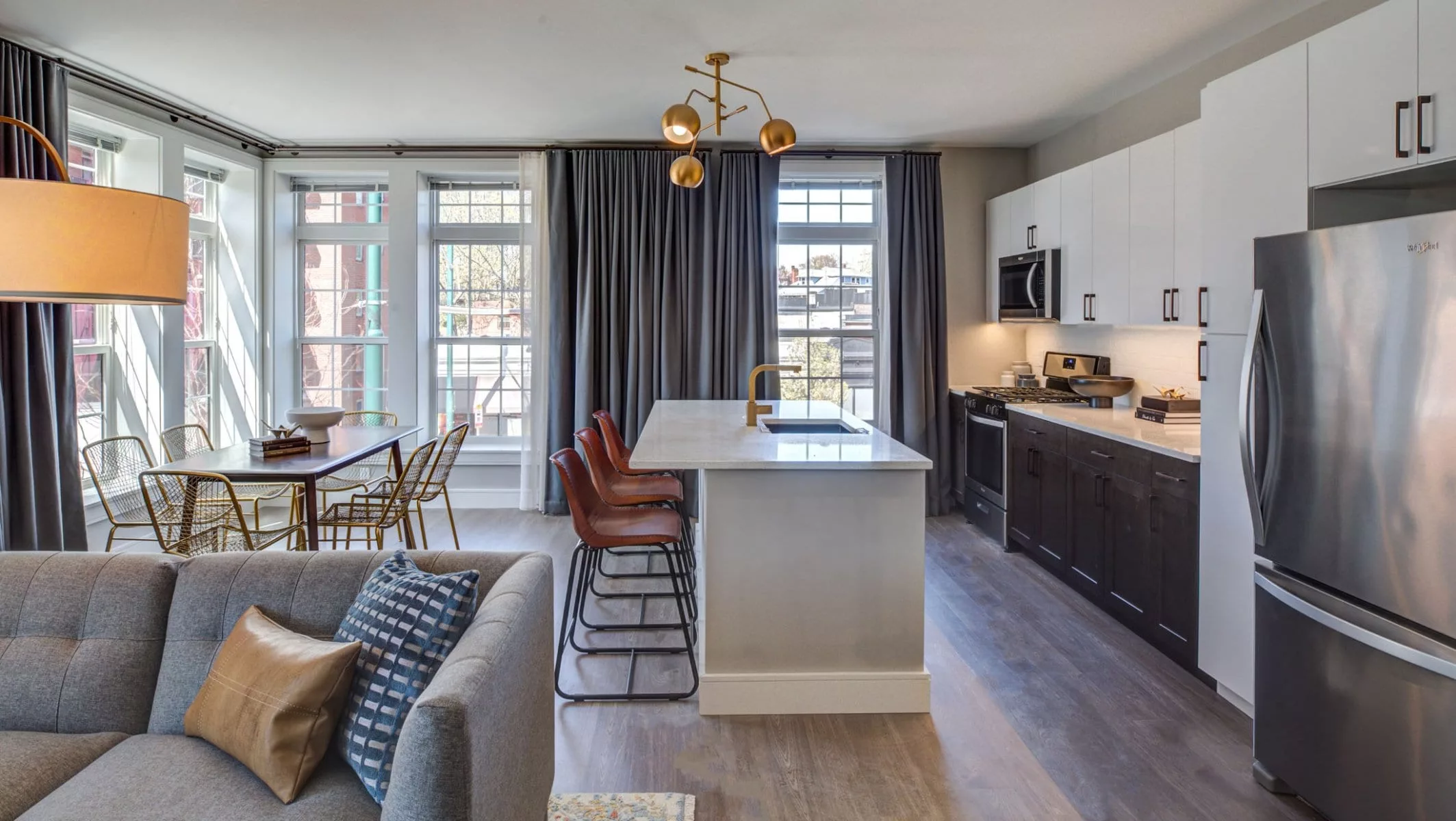 Large living room, dining room, and kitchen area in an apartment at The Bradford, with several bright windows, wood-style flooring, stainless steel appliances, and quartz countertops